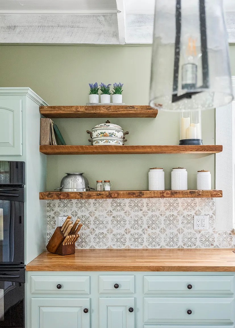 Sage green kitchen wall with three wooden floating shelves styled with dishes and decor
