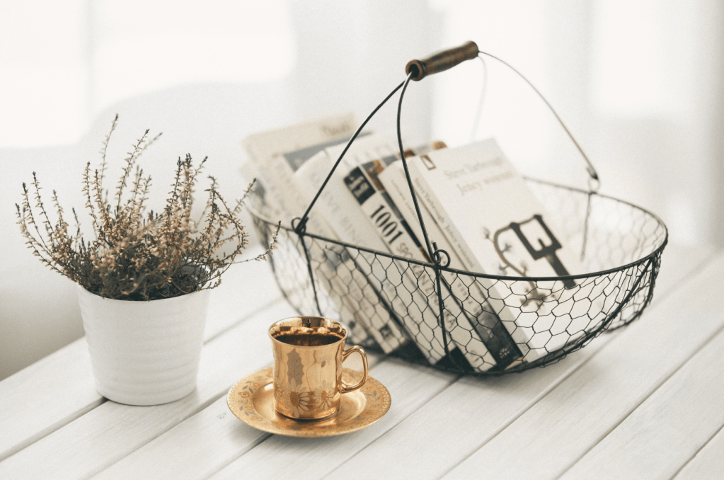 Wire basket with books sitting on a bright coffee table next to a gold coffeecup and saucer and a plant